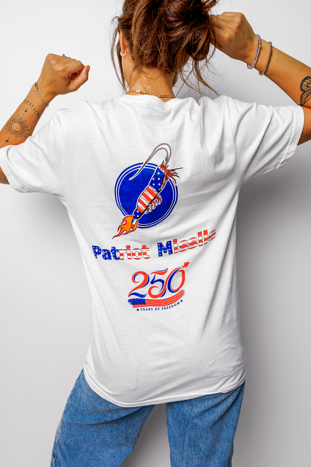 A woman shown from behind wearing a white graphic t-shirt with a patriotic missile design and blue jeans, posing with her fists raised against a light gray background.