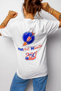 A woman shown from behind wearing a white graphic t-shirt with a patriotic missile design and blue jeans, posing with her fists raised against a light gray background.