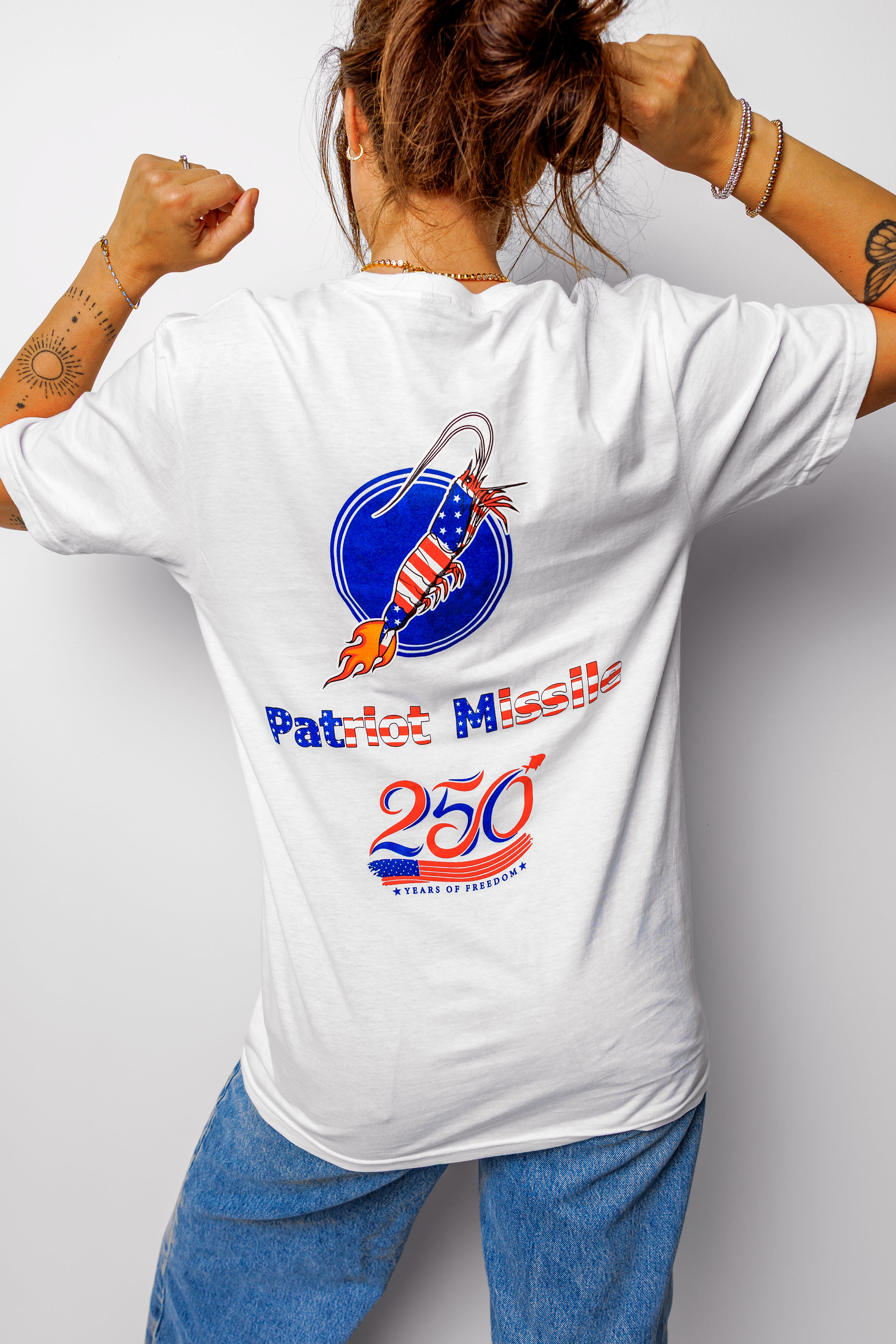A woman shown from behind wearing a white graphic t-shirt with a patriotic missile design and blue jeans, posing with her fists raised against a light gray background.