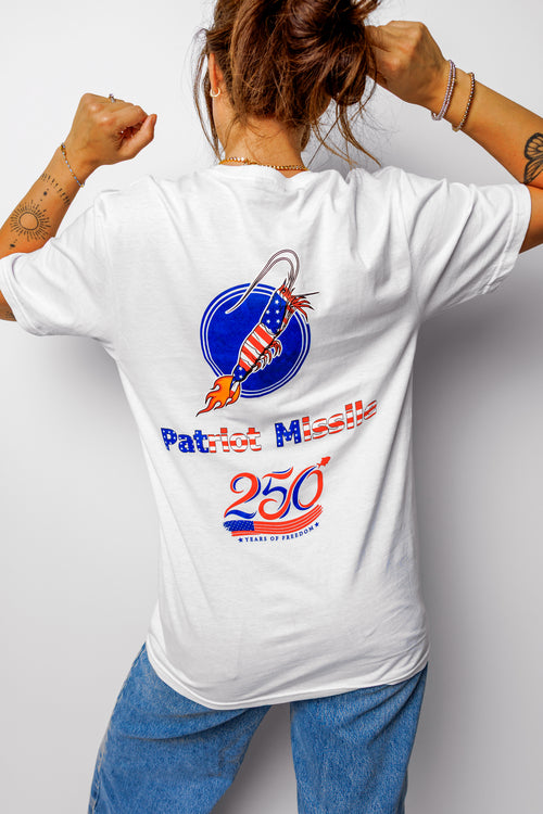 A woman shown from behind wearing a white graphic t-shirt with a patriotic missile design and blue jeans, posing with her fists raised against a light gray background.