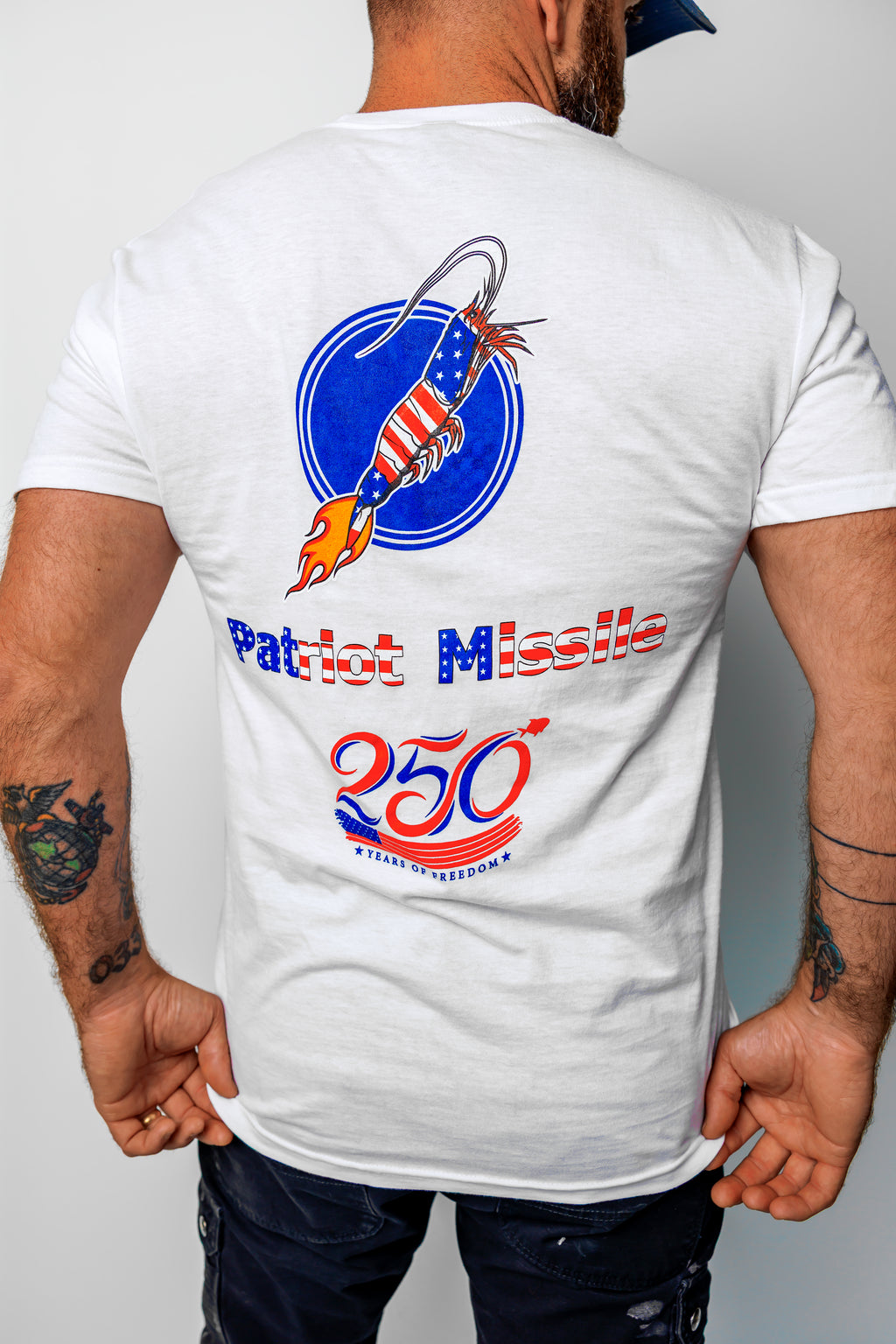 A man wearing a white t-shirt, viewed from the back. The shirt features a large graphic print of a patriotic missile inside a blue circle, with the text 'Patriot Missile' and '250th' in red and blue.